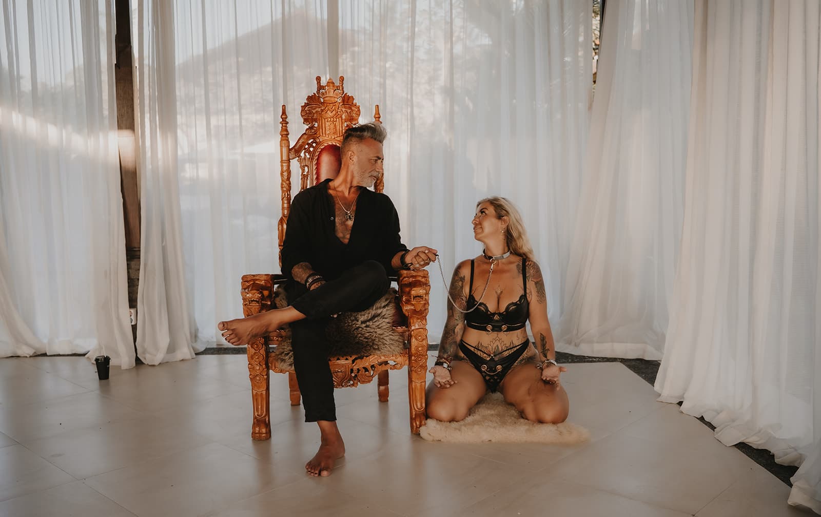 Rex seated on a carved throne holding a leash, with Rebelle kneeling beside him in a collar, looking up at him